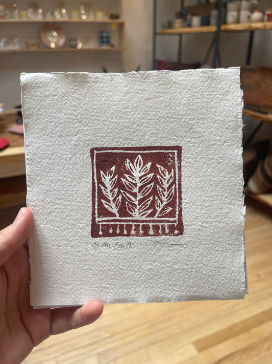 A hand holding a 6 by 6 inch block print with a dark red botanical design, signed and crafted by Rebecca Kunz.